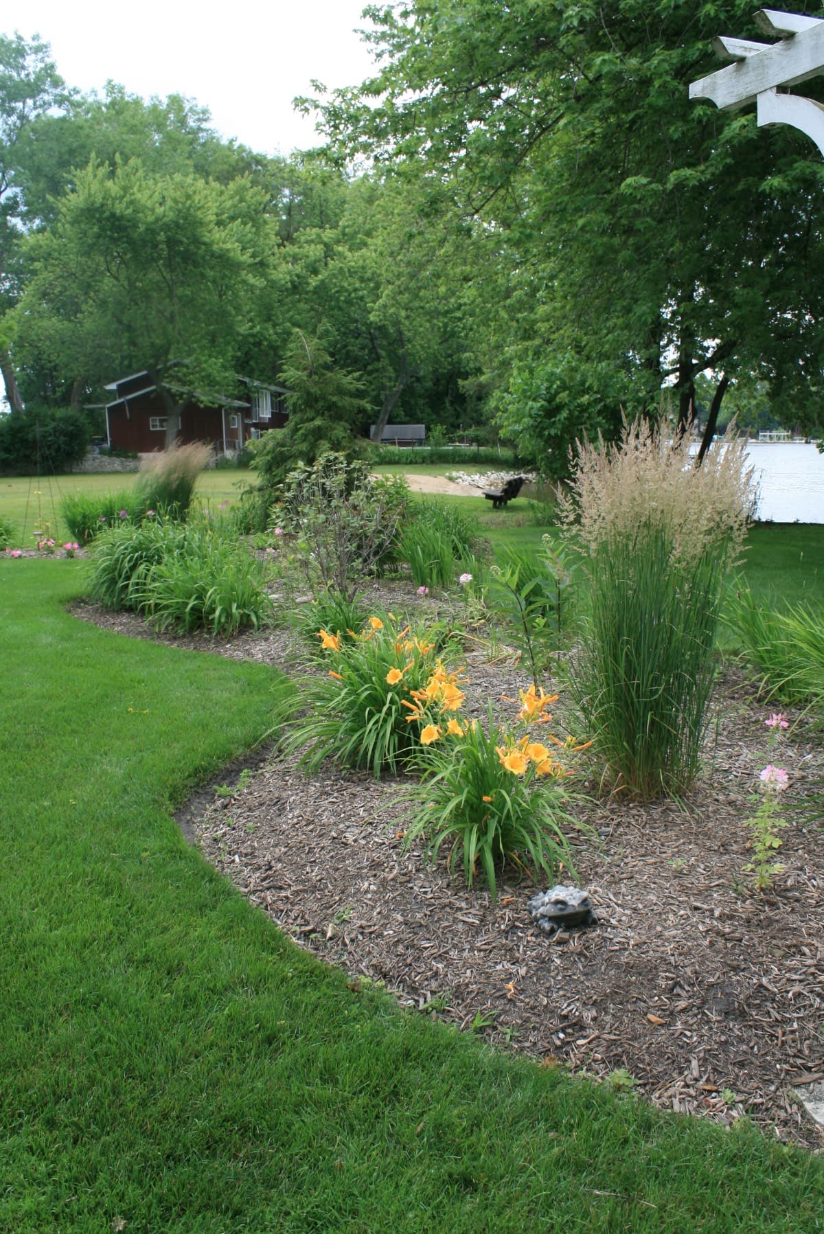 A garden bed with young plants and small white plant markers, surrounded by mulch. Nearby, there is a weathered wooden bench and a large planter with yellow-green foliage. Grass and trees are in the background.