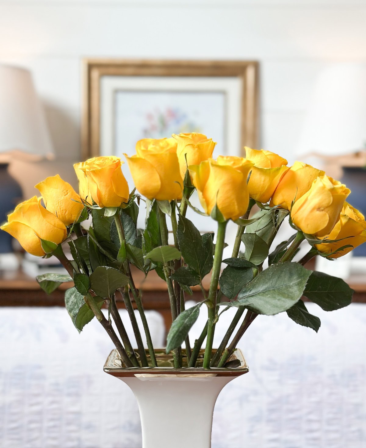 A vase filled with a bouquet of yellow roses, set on a table in a well-lit room with framed art and two lamps in the background.