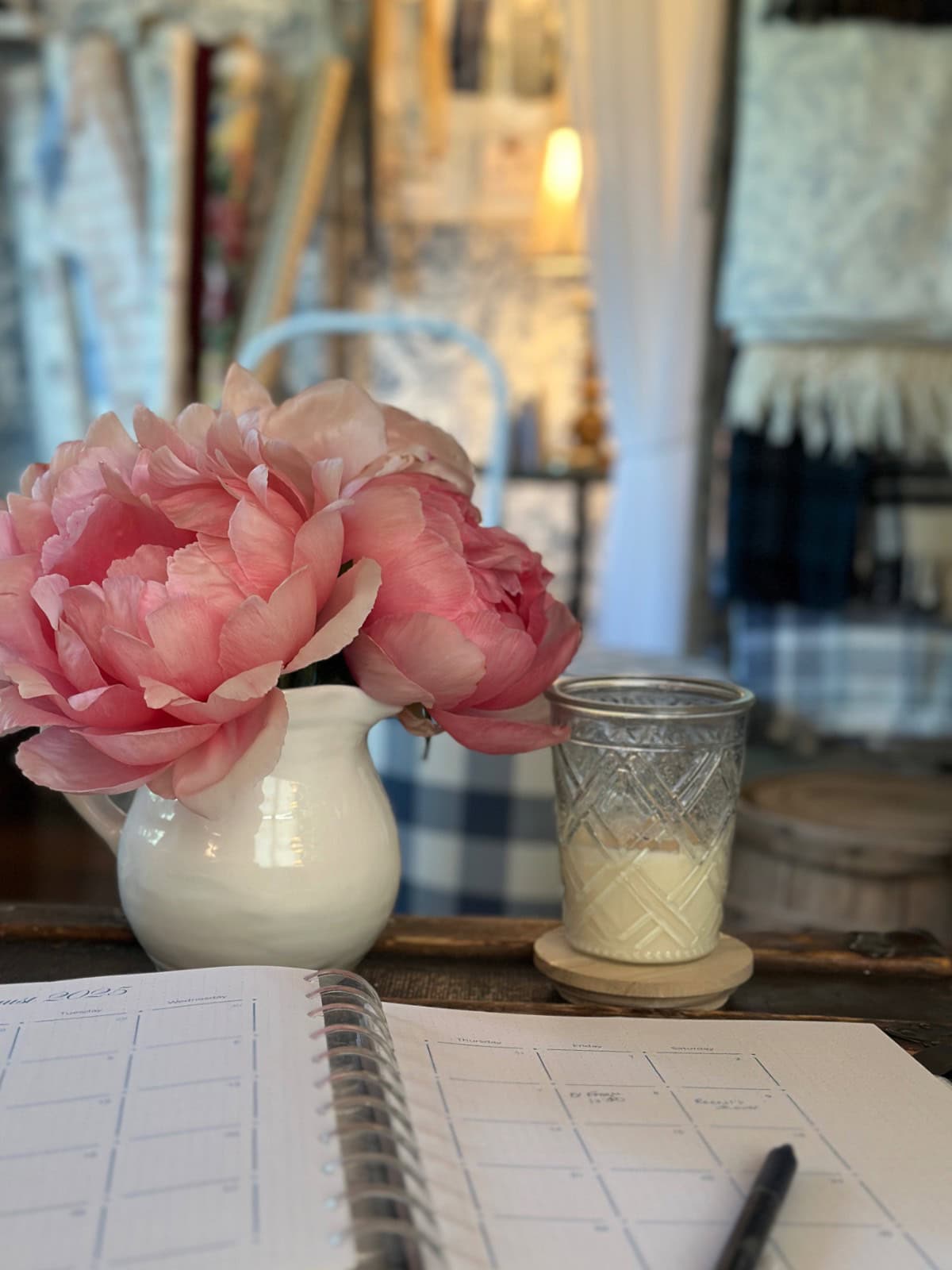 A white pitcher with pink peonies and a jar candle sit on a table next to an open planner and pen, creating a cozy scene with the latest home decor softly blurred in the background.