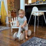 A toddler in a gray "Chill Mood" shirt rides the latest white balance bike indoors on a wooden floor, with a high chair, a white chair, and household items visible in the background.