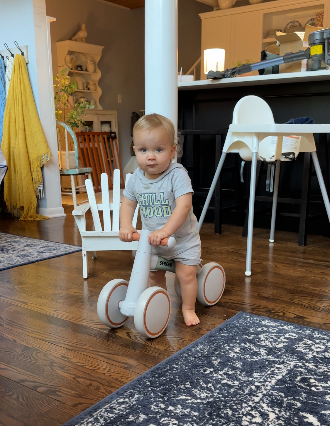 A toddler wearing a gray "Chill Mood" shirt stands on a wooden floor, holding onto the latest white balance bike, with a high chair, chair, and kitchen area visible in the background.