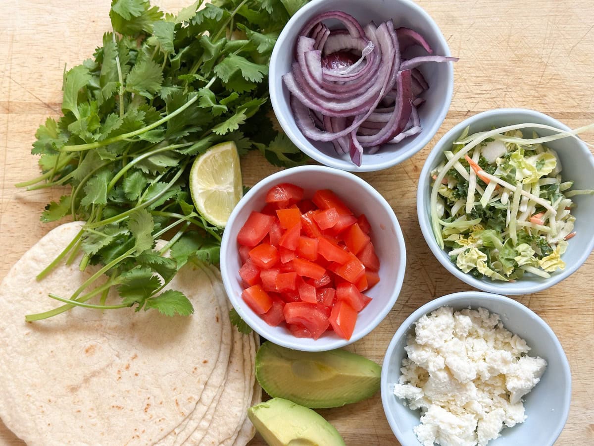 A wooden board with tortillas, fresh cilantro, lime wedges, sliced avocado, bowls of red onion, shredded cabbage mix, diced tomato, and crumbled cheese, arranged for taco preparation.