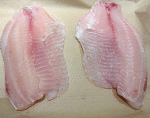 Two raw white fish fillets placed side by side on a sheet of brown parchment paper, showing their translucent texture and pinkish lines.