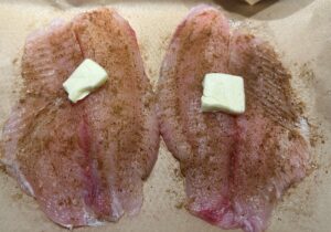 Two raw fish fillets on parchment paper, sprinkled with seasoning and each topped with a pat of butter, ready for cooking.