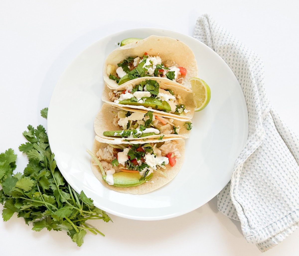 Three soft tacos filled with baked fish, avocado slices, slaw, fresh cilantro, crumbled cheese, and drizzled with spicy cream sauce, served on a white plate with cilantro garnish.