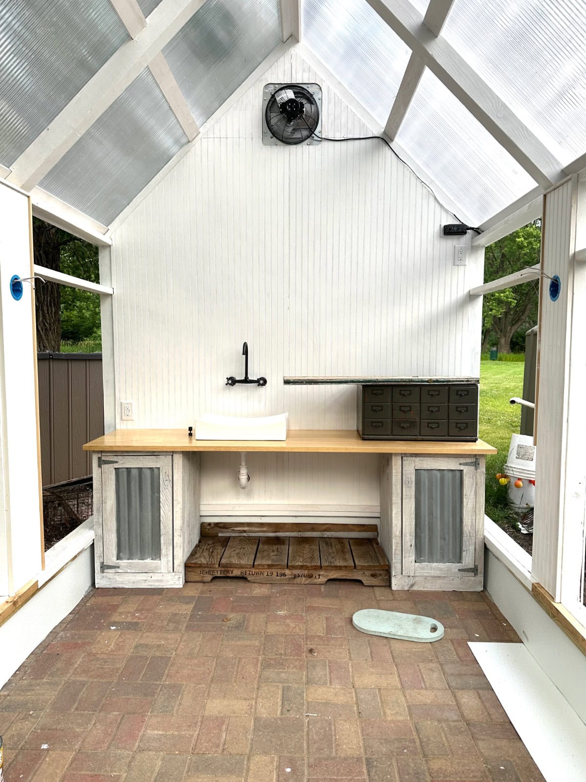 A latest potting bench with a sink, faucet, rustic cabinets, and drawers sits inside a bright greenhouse with a clear roof and brick floor. A garden is visible through the glass walls.