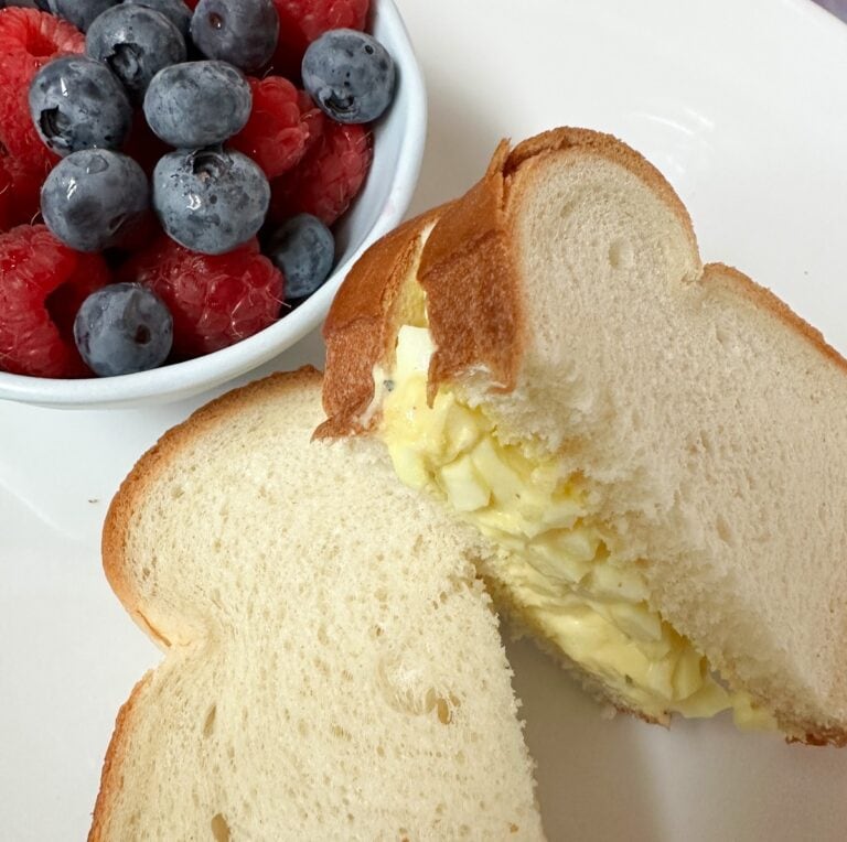 A plate with an egg salad sandwich on white bread, next to a small bowl filled with fresh blueberries and raspberries.
