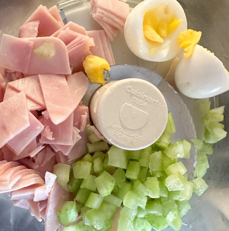 Chopped ham, celery, and hard-boiled eggs in a food processor bowl, ready to be mixed.