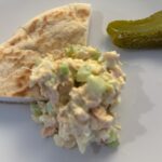 A serving of chicken salad with diced celery on a white plate, accompanied by a wedge of pita bread and two small pickles.