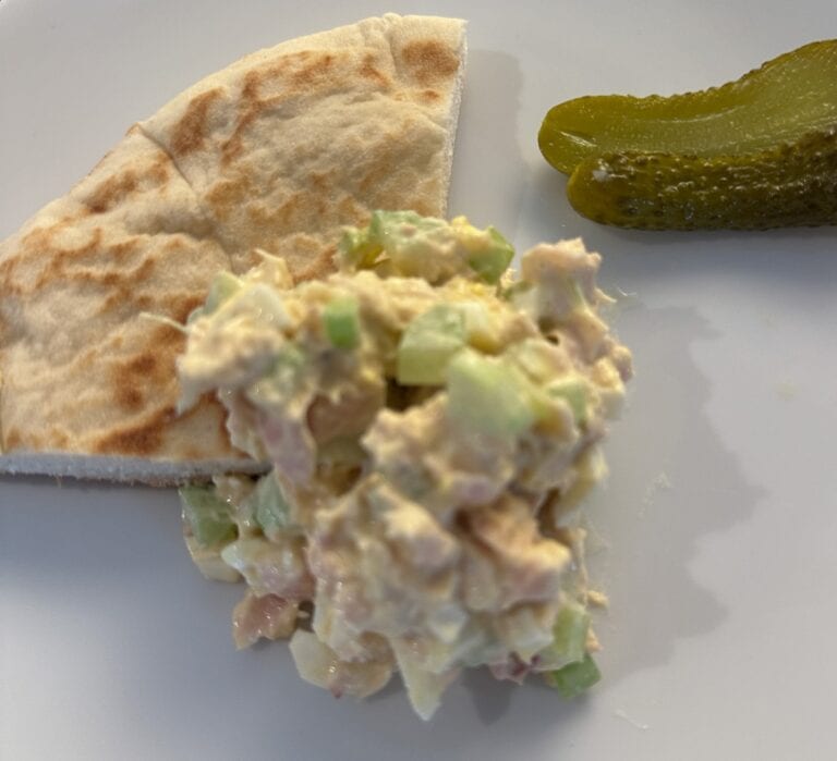 A serving of chicken salad with diced celery on a white plate, accompanied by a wedge of pita bread and two small pickles.