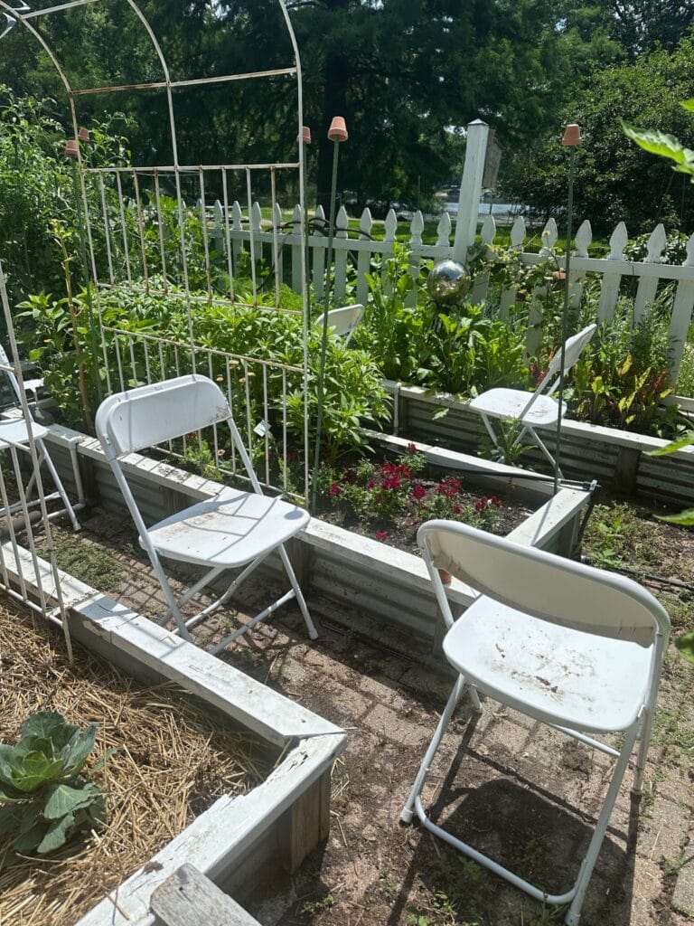 Latest white folding chairs are arranged among raised garden beds filled with green plants and flowers, with a white picket fence and trellis in the background on a sunny day.