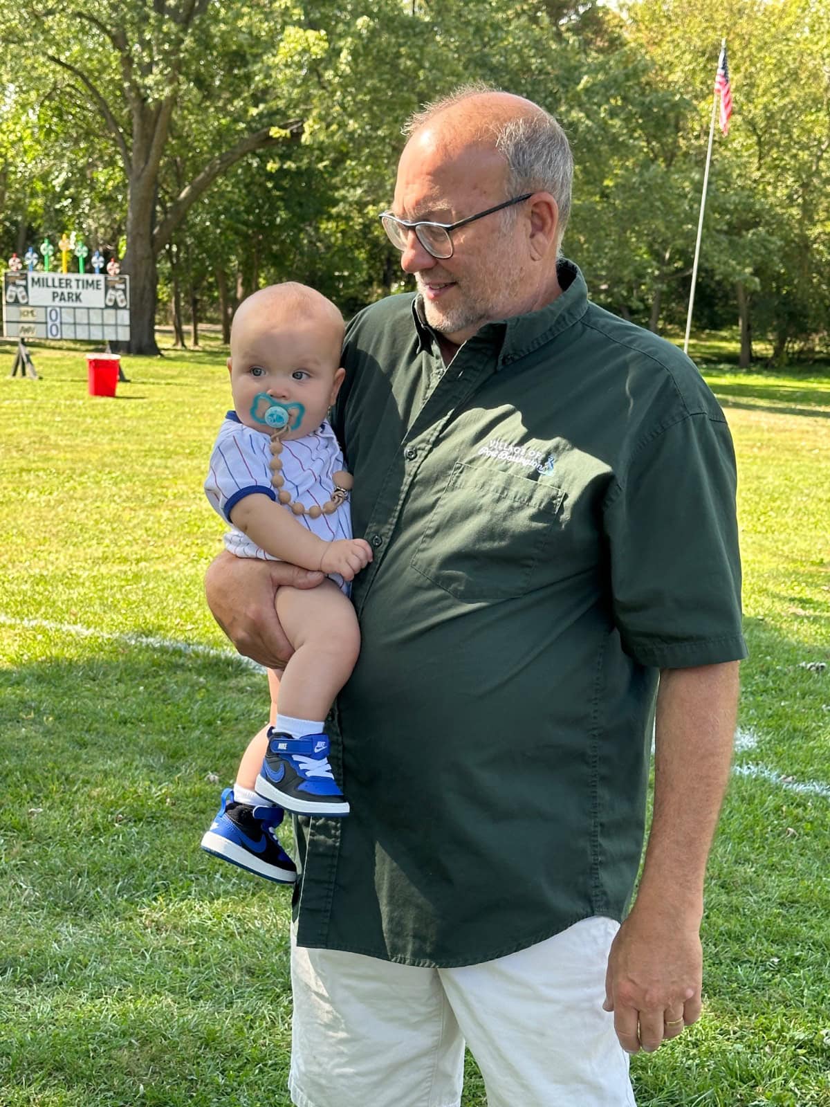 An older man wearing glasses and a green shirt holds a baby dressed in a striped outfit and blue shoes. They are standing on grass in a park with trees, a scoreboard, and an American flag in the background.