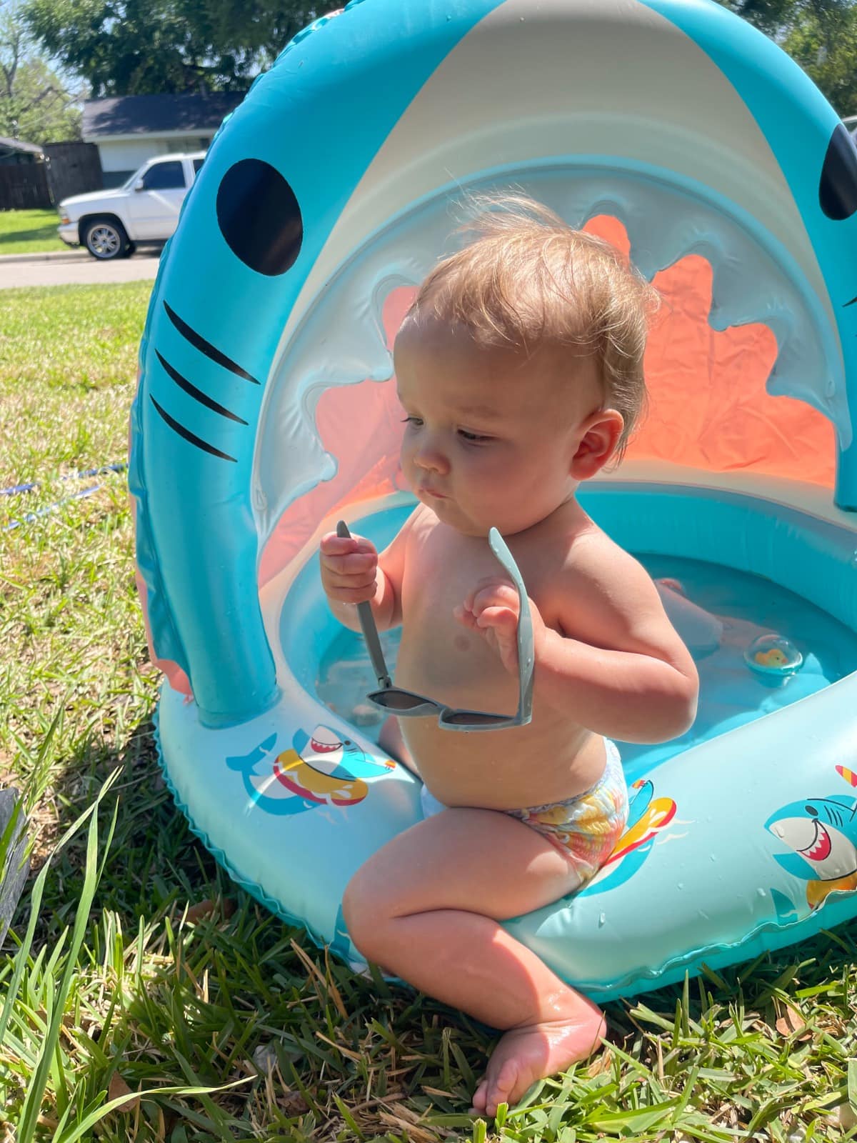 A baby in a diaper sits in a small, shark-shaped inflatable pool on grass, holding sunglasses and looking to the side on a sunny day.