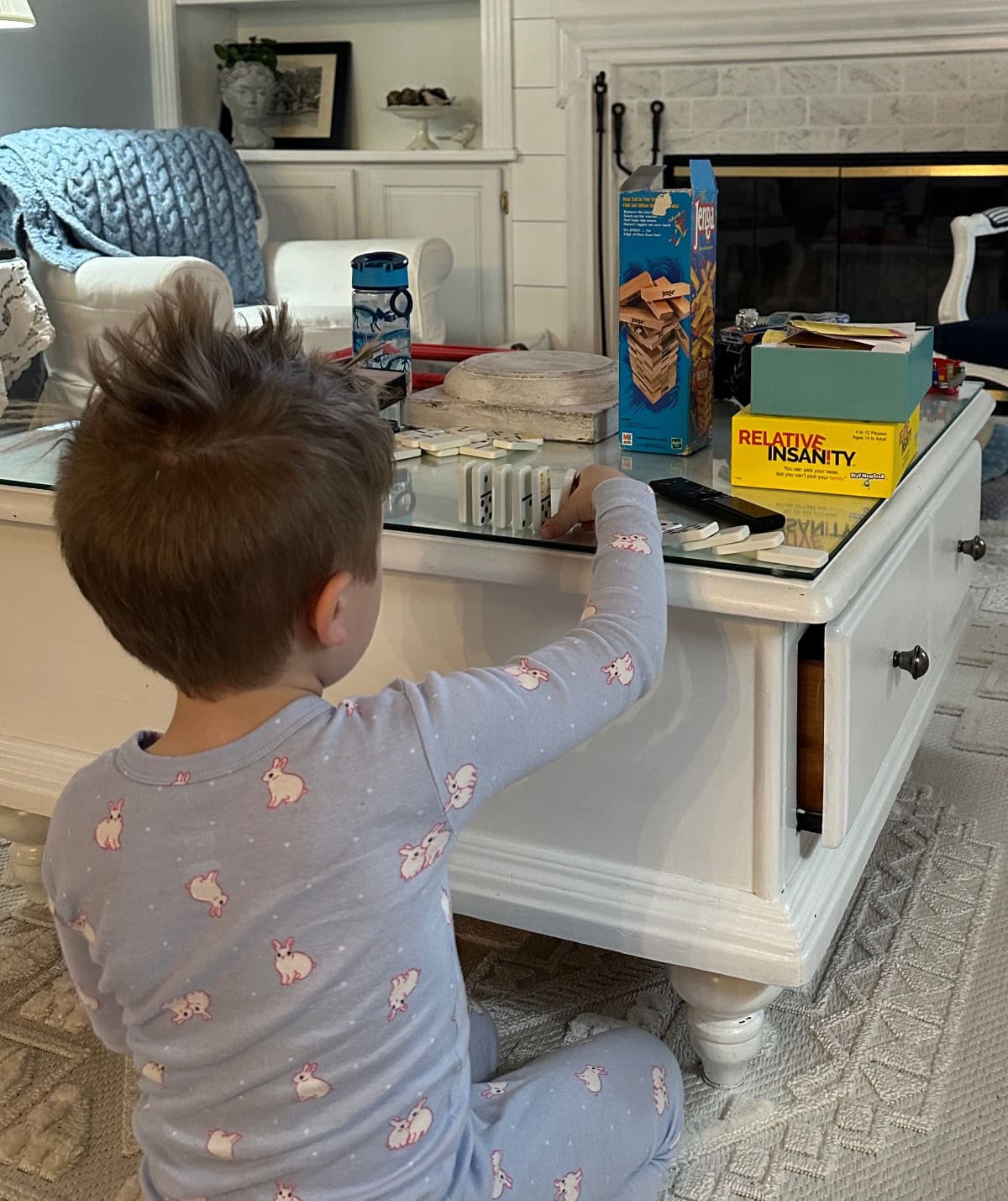 A young child in bunny pajamas sits on the floor, reaching for items on a coffee table with games, snacks, and cards—perfect for long distance grandparenting moments shared over video calls in a cozy living room.