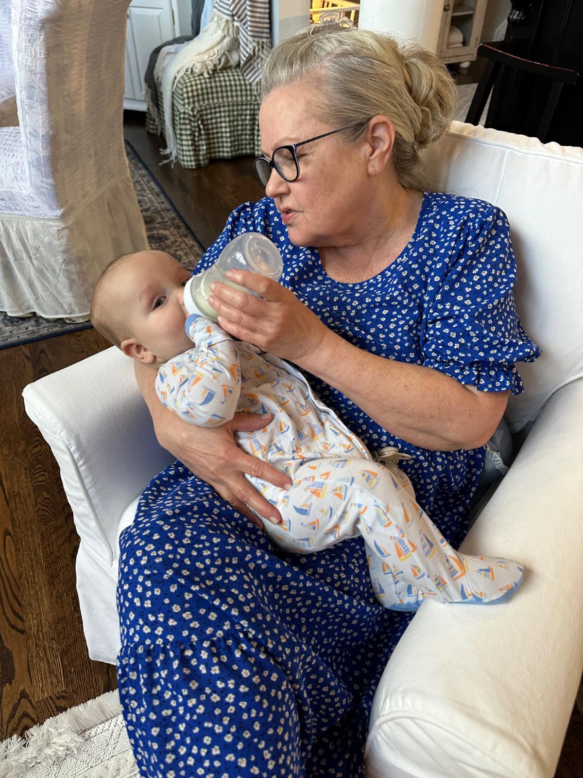 An older woman in glasses and a blue floral dress sits in a white armchair, feeding a baby with a bottle. The baby, dressed in a patterned onesie, rests comfortably on her lap. The setting appears to be a cozy living room.