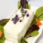 A rectangular cake with white frosting is garnished with purple flowers and surrounded by green and dark red leafy vegetables on a white platter. Orange flowers also decorate the platter.