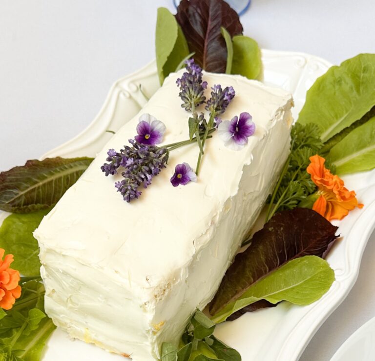 A rectangular cake with white frosting is garnished with purple flowers and surrounded by green and dark red leafy vegetables on a white platter. Orange flowers also decorate the platter.