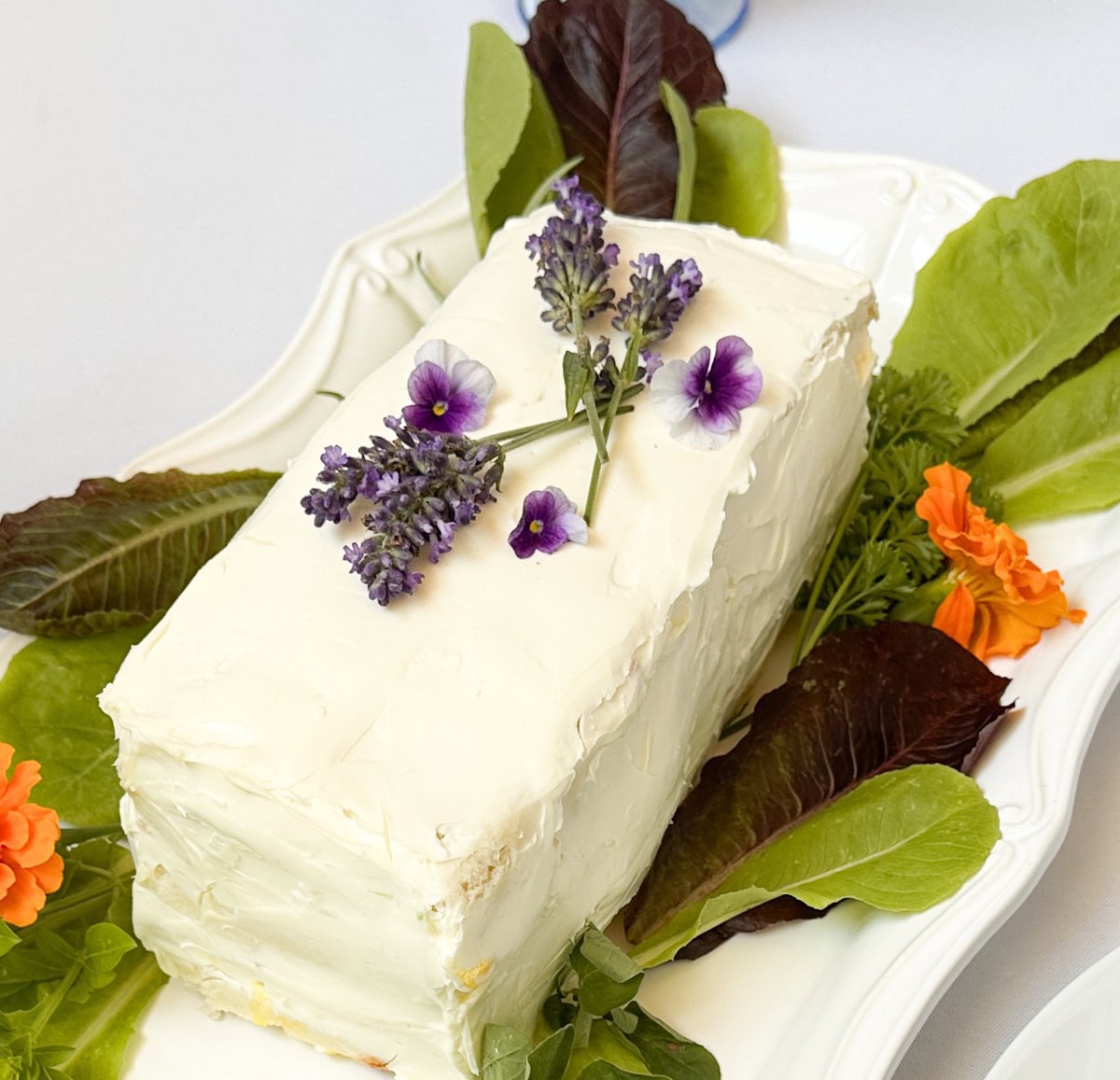 A rectangular cake with white frosting is garnished with purple flowers and surrounded by green and dark red leafy vegetables on a white platter. Orange flowers also decorate the platter.