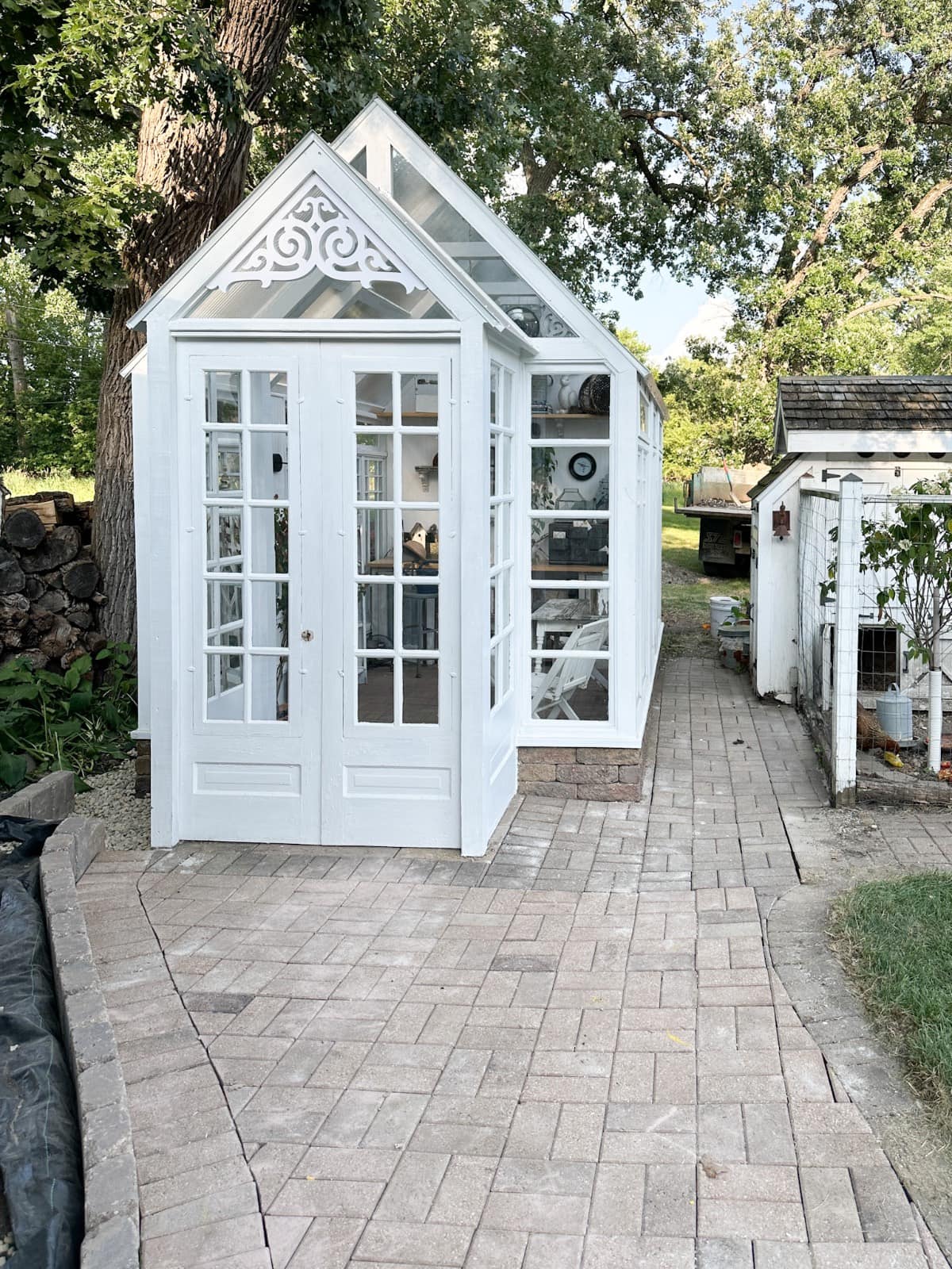 The latest addition to the garden, a small white greenhouse with glass windows and ornate trim, sits on a paved walkway, surrounded by trees, a woodpile, and a nearby small white shed or chicken coop.