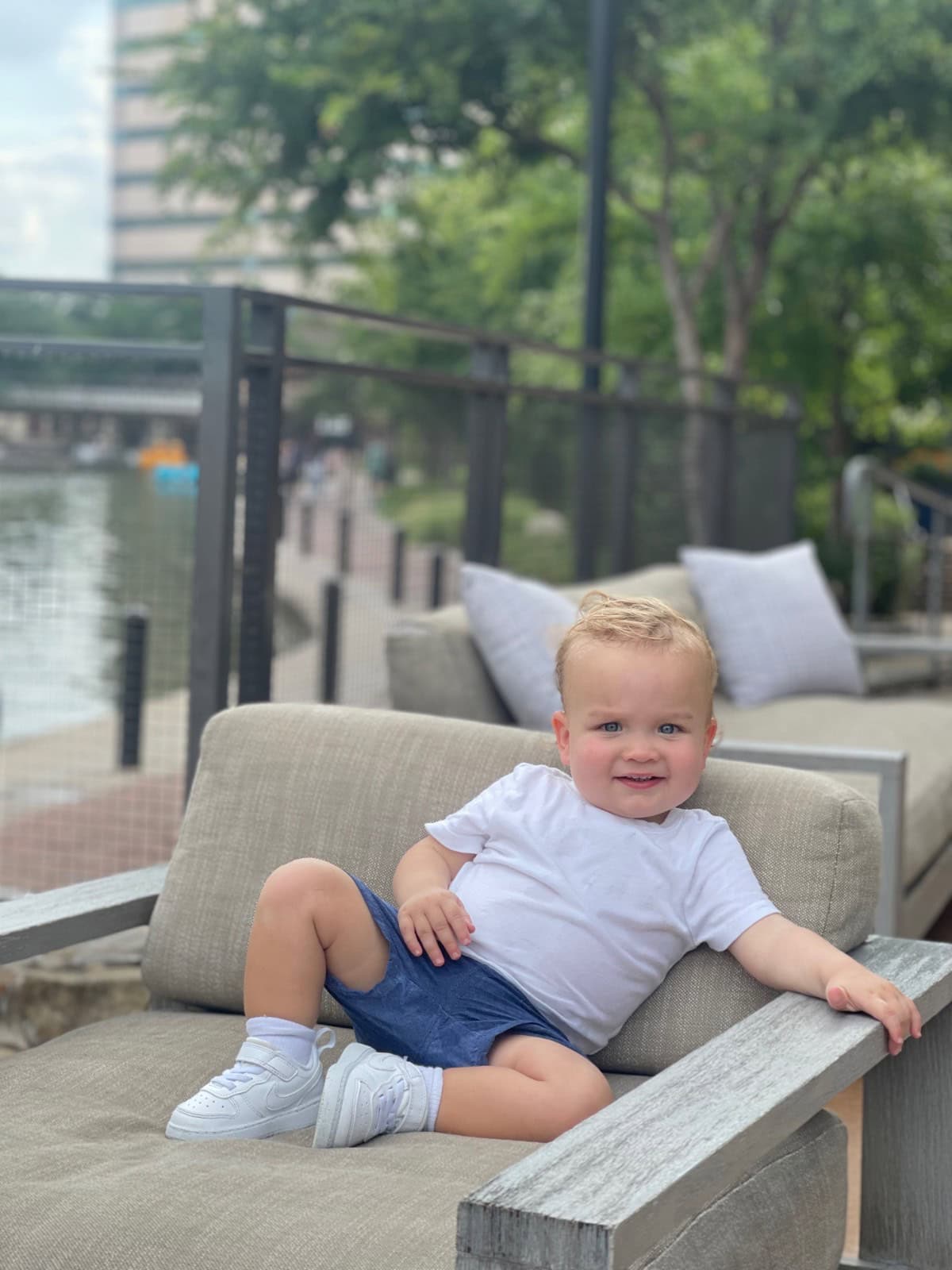 A smiling toddler in a white t-shirt, blue shorts, and white sneakers sits relaxed on an outdoor cushioned lounge chair by the waterfront after a fun Saturday shopping trip, with trees and buildings visible in the background.