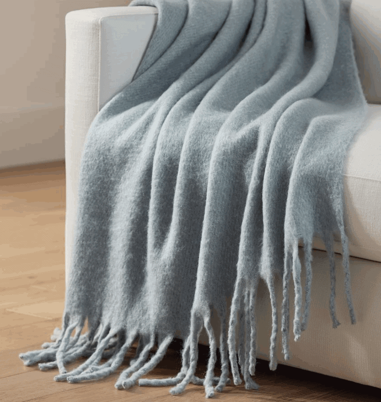 A soft, light blue throw blanket with long fringe drapes over the arm of a white sofa, resting on a wooden floor in a cozy, sunlit room—perfect for your next Saturday Shopping find.