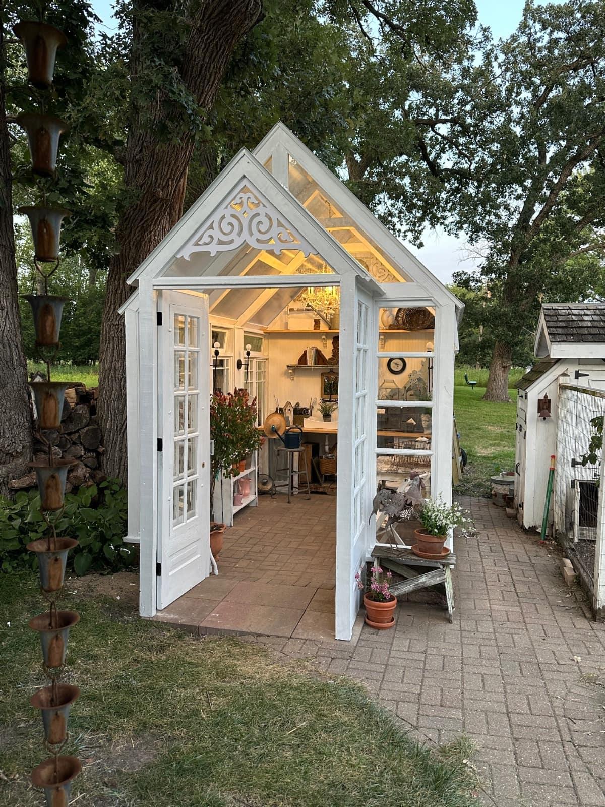 A small, white greenhouse with glass walls and doors stands open in a garden. Inside, shelves display the latest plants and gardening tools. Potted plants and a bench sit outside on a paved path, surrounded by trees and greenery.