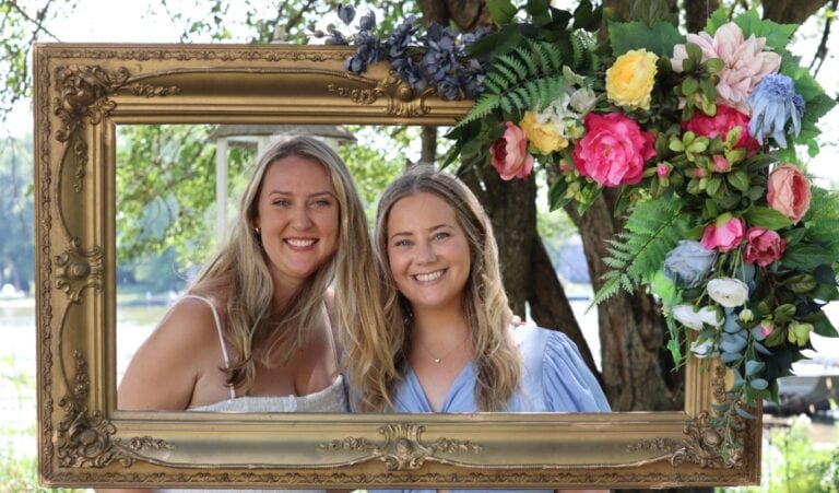 Two women smiling and posing together outdoors, framed by a vintage frame adorned with a faux flower swag and lush greenery, with trees and a lake in the background.