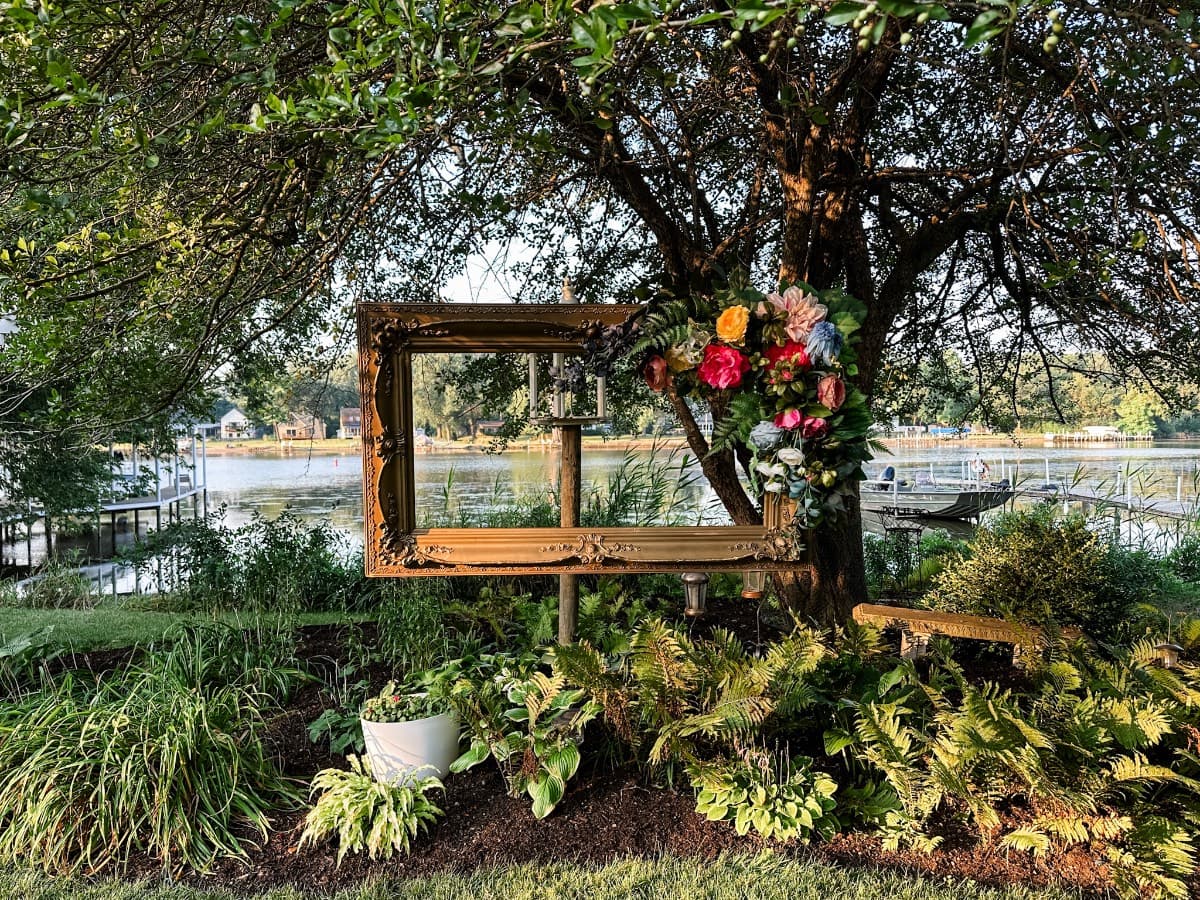 A large empty vintage frame adorned with a faux flower swag is displayed outdoors under a leafy tree, with a lake, docks, and houses in the background, surrounded by lush green plants and landscaping.