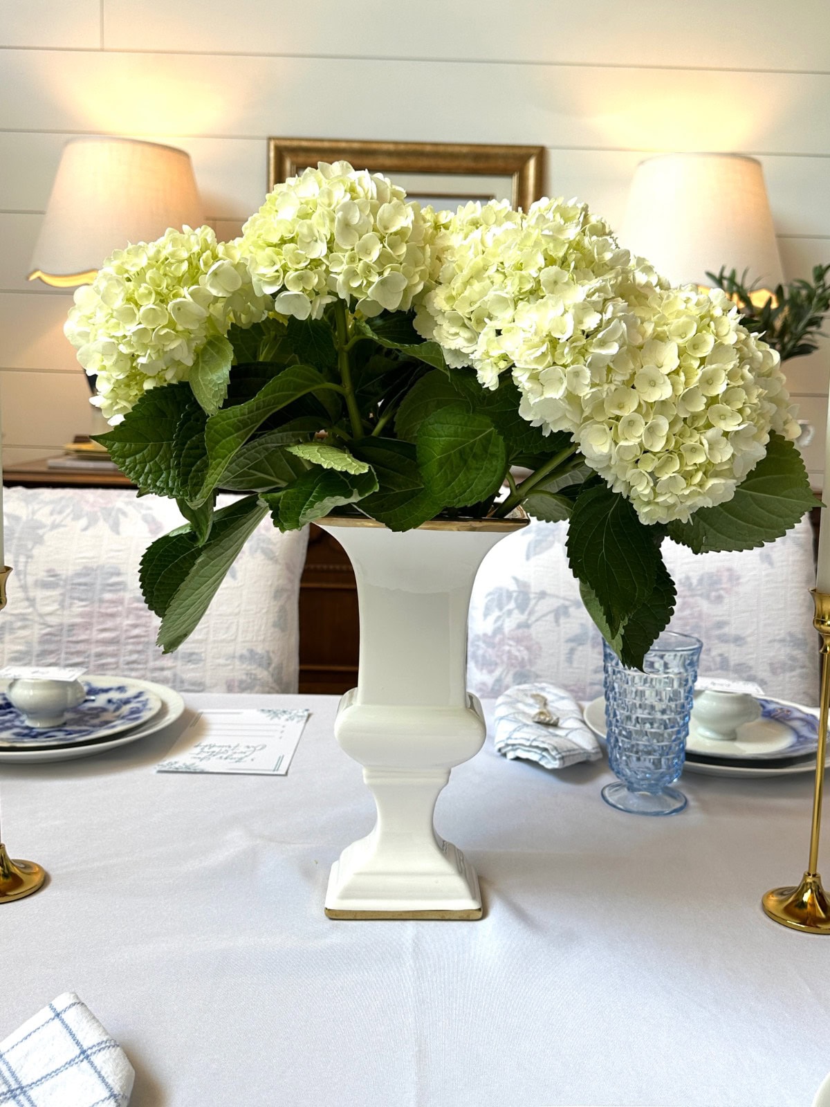A white vase with large white hydrangea flowers sits on a neatly set dining table, reminiscent of shopping with friends for elegant gold candlesticks, patterned plates, and blue glassware in a warmly lit dining room.