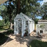 A small white greenhouse with glass windows and a pitched roof sits on a brick patio in a sunny backyard, surrounded by trees and plants. A chicken coop is visible nearby.