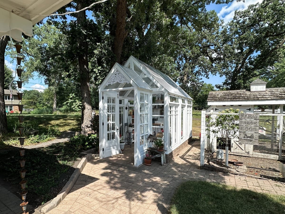 A small white greenhouse with glass windows and a pitched roof sits on a brick patio in a sunny backyard, surrounded by trees and plants. A chicken coop is visible nearby.