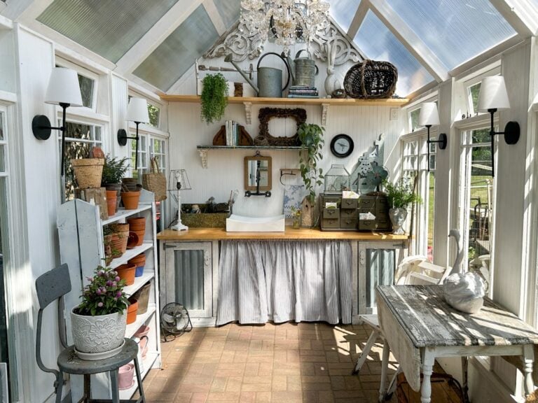 Bright, sunlit greenhouse with potted plants on shelves, a rustic sink, decorative accents, and gardening supplies. Windows line the walls and roof, filling the cozy space with natural light.