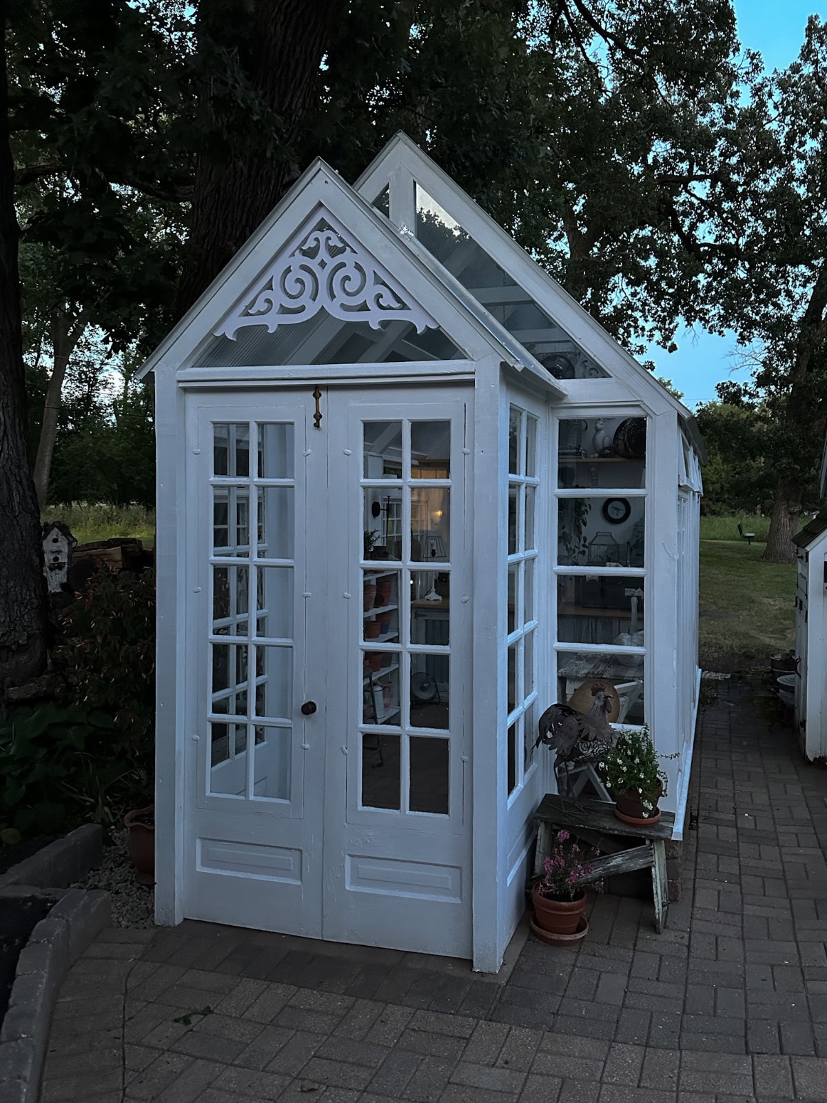 A small, white greenhouse with glass doors and windows stands on a paved path, surrounded by trees and plants. Decorative trim adorns the peak, and potted plants sit near the entrance. Shelves are visible inside.