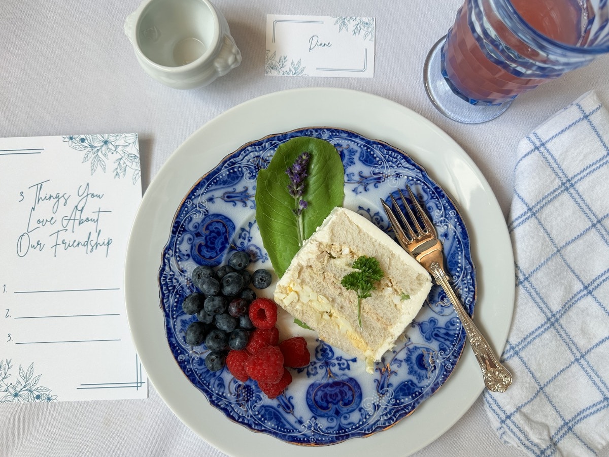 A blue and white plate holds a slice of dessert, blueberries, raspberries, and a leaf garnish. A gold fork, a glass of pink drink, note cards, and a blue-check napkin are arranged on a white tablecloth.