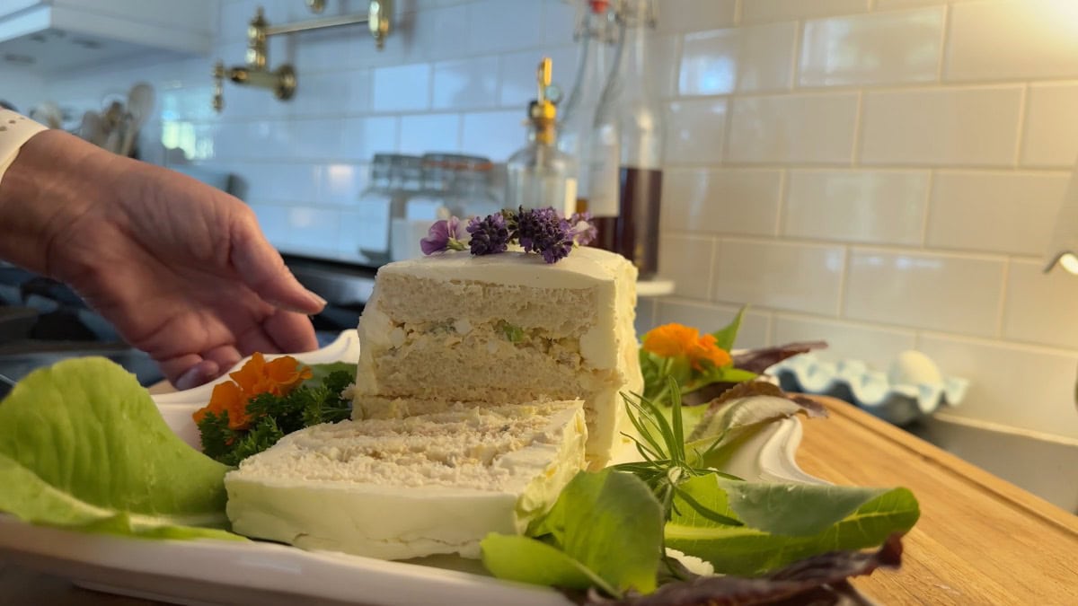 A hand is placing a plate with a layered savory cake, decorated with lettuce, herbs, and edible flowers, on a wooden surface in a bright kitchen with white tile walls.