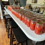 Rows of glass jars filled with red tomato sauce are lined up on a long white kitchen counter, with several black stools underneath. The kitchen has wooden floors and white cabinets.