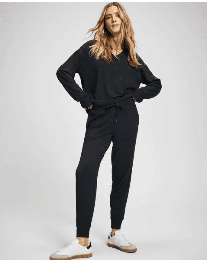 A woman stands confidently, ready for Saturday shopping in a black long-sleeve pullover and matching jogger pants with white and beige sneakers. Her hands are in her pockets and she smiles slightly against a plain, light background.