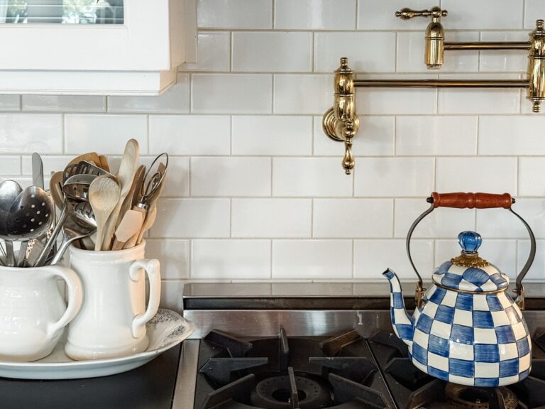 A blue and white checkered teapot sits on a stove next to two white ceramic containers holding various kitchen utensils—perfect inspiration for Fall Home Organizing, all set against a chic white subway tile backsplash and brass pot filler above.