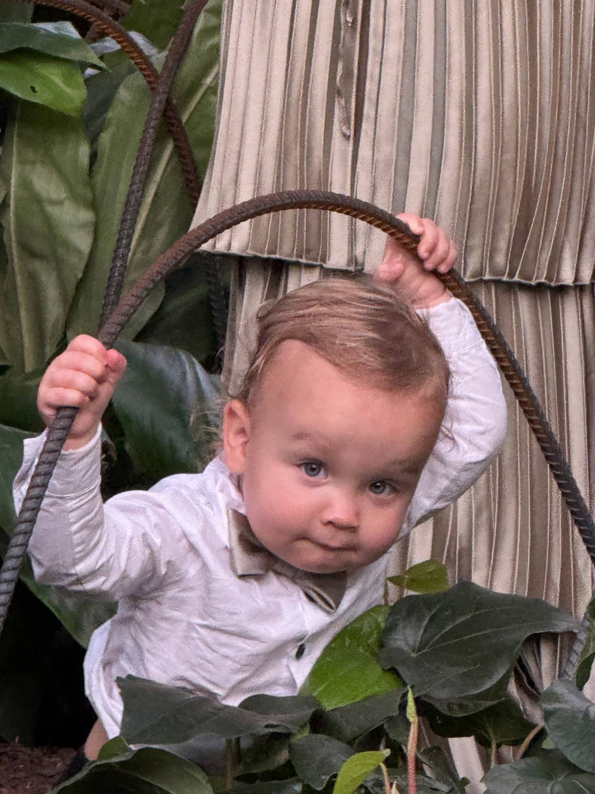 A young child in a white shirt and bow tie holds onto a curved metal rod, standing among green leaves. Behind the child, part of a person in a pleated beige skirt showcases the latest in chic, understated style.