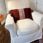 A white upholstered armchair with a DIY Sweater Pillow featuring a red and white geometric pattern and tassels sits on a wooden floor next to a round side table in a cozy living room.