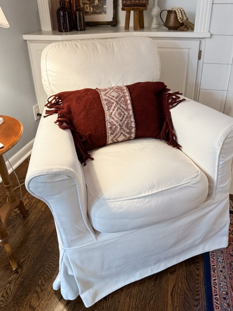 A white upholstered armchair with a DIY Sweater Pillow featuring a red and white geometric pattern and tassels sits on a wooden floor next to a round side table in a cozy living room.