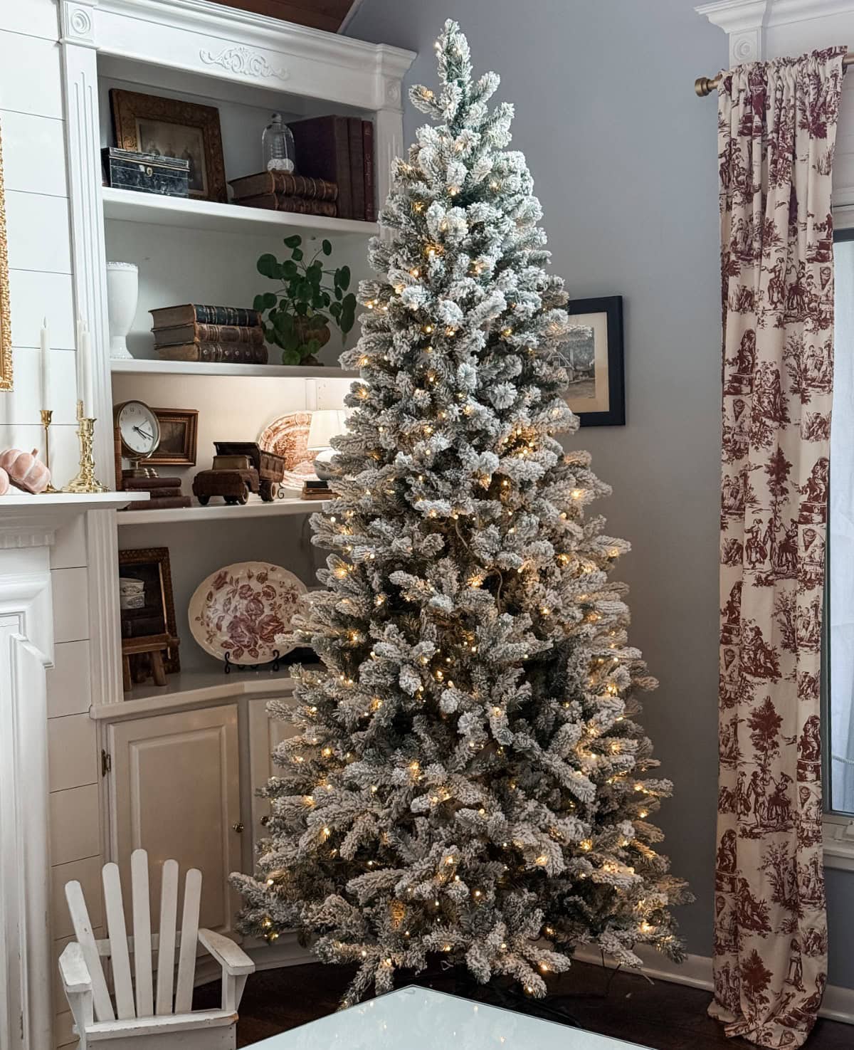 A tall, flocked Christmas tree with white lights stands in a cozy living room corner, surrounded by shelves with books, vintage decor, and a window with patterned curtains.