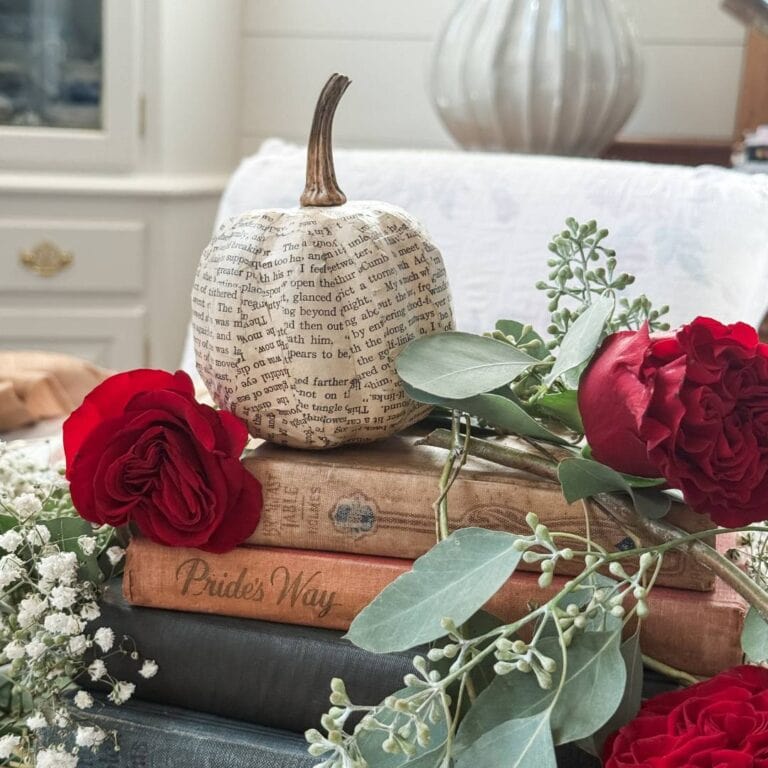A small pumpkin covered in book pages sits on a stack of vintage books. Red roses, greenery, and a wine bottle candle are draped around the books, creating a cozy, literary-themed arrangement on a soft, neutral background.