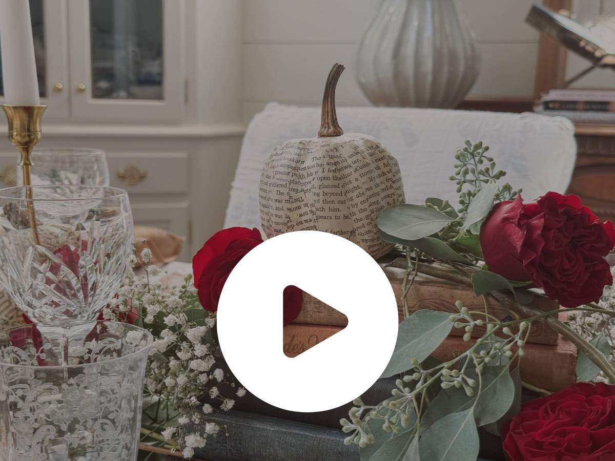 A decorated table with crystal glasses, red roses, greenery, and a book page pumpkin sits in a bright, elegant room. A large play button icon is centered over the image.