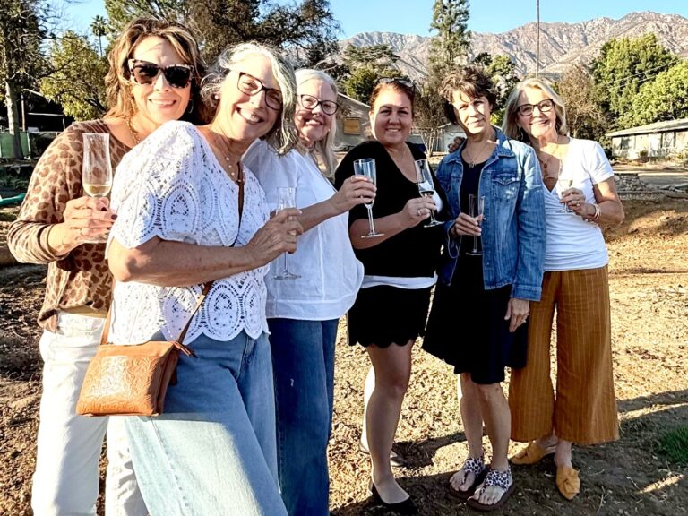 Six women stand outdoors smiling, holding champagne glasses. The group, possibly celebrating a podcast milestone, is casually dressed with trees and mountains visible in the background on a sunny day.