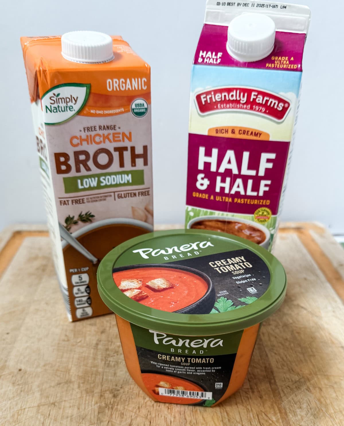 A carton of Simply Nature organic low-sodium chicken broth, a carton of Friendly Farms half & half, and a container of Panera Bread creamy tomato soup sit on a wooden surface.