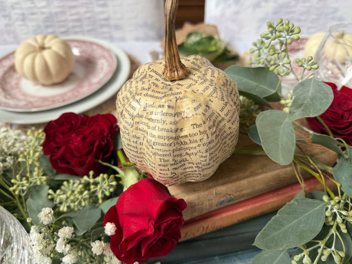 A small book page pumpkin sits on stacked books, surrounded by red roses, eucalyptus leaves, and baby's breath, with a blurred plate and another pumpkin in the background.