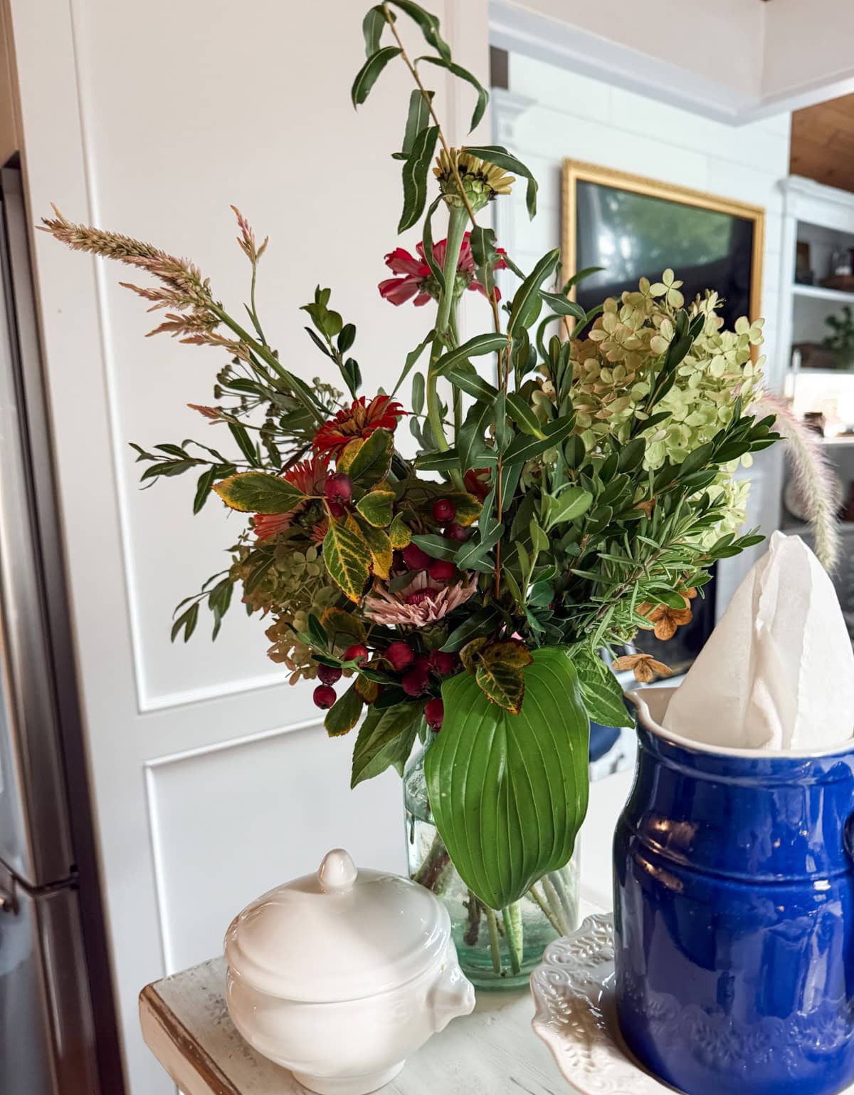 A glass vase filled with green leaves, red berries, and white flowers sits on a white table next to book page pumpkins, a blue container holding napkins, and a small white ceramic sugar bowl with a lid.