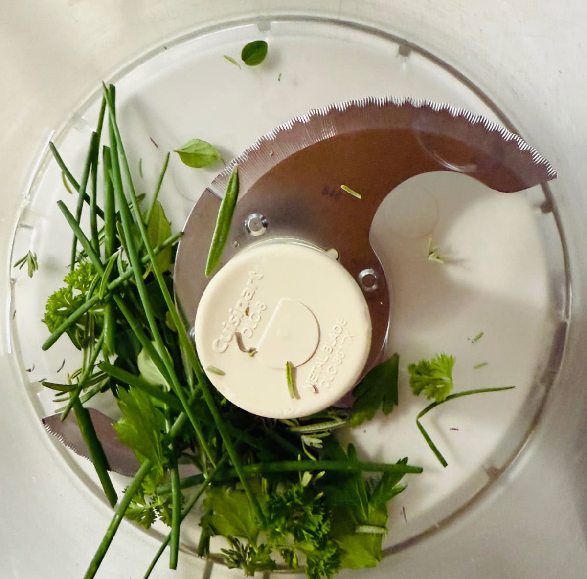 Fresh herbs, including chives and parsley, are placed inside a food processor bowl with a sharp metal blade attachment, ready to be chopped.