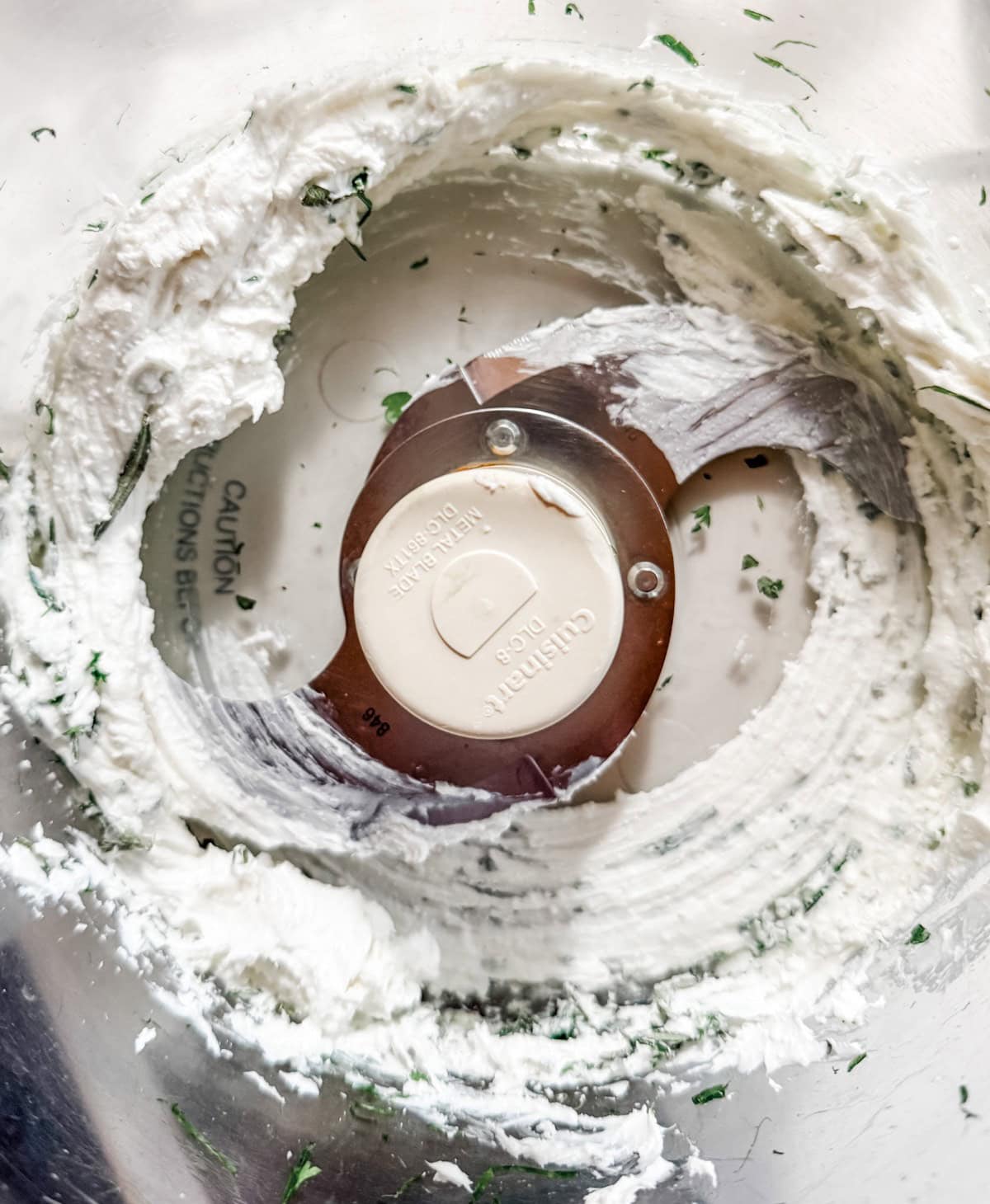 Close-up of a food processor bowl with creamy, white mixture and green herb bits stuck to the sides around the metal blade.